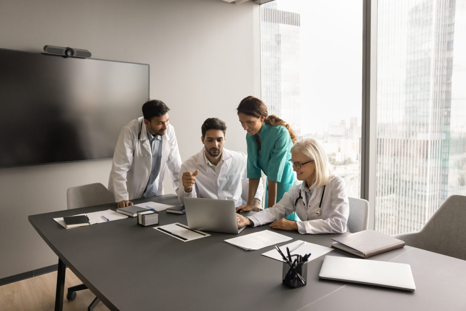 Team of medical colleagues using laptop for Internet communication Four professionals, including a medical billing specialist, gather in a conference room to discuss a project around a laptop. Papers and notebooks are scattered on the table while large windows reveal the bustling cityscape in the background.